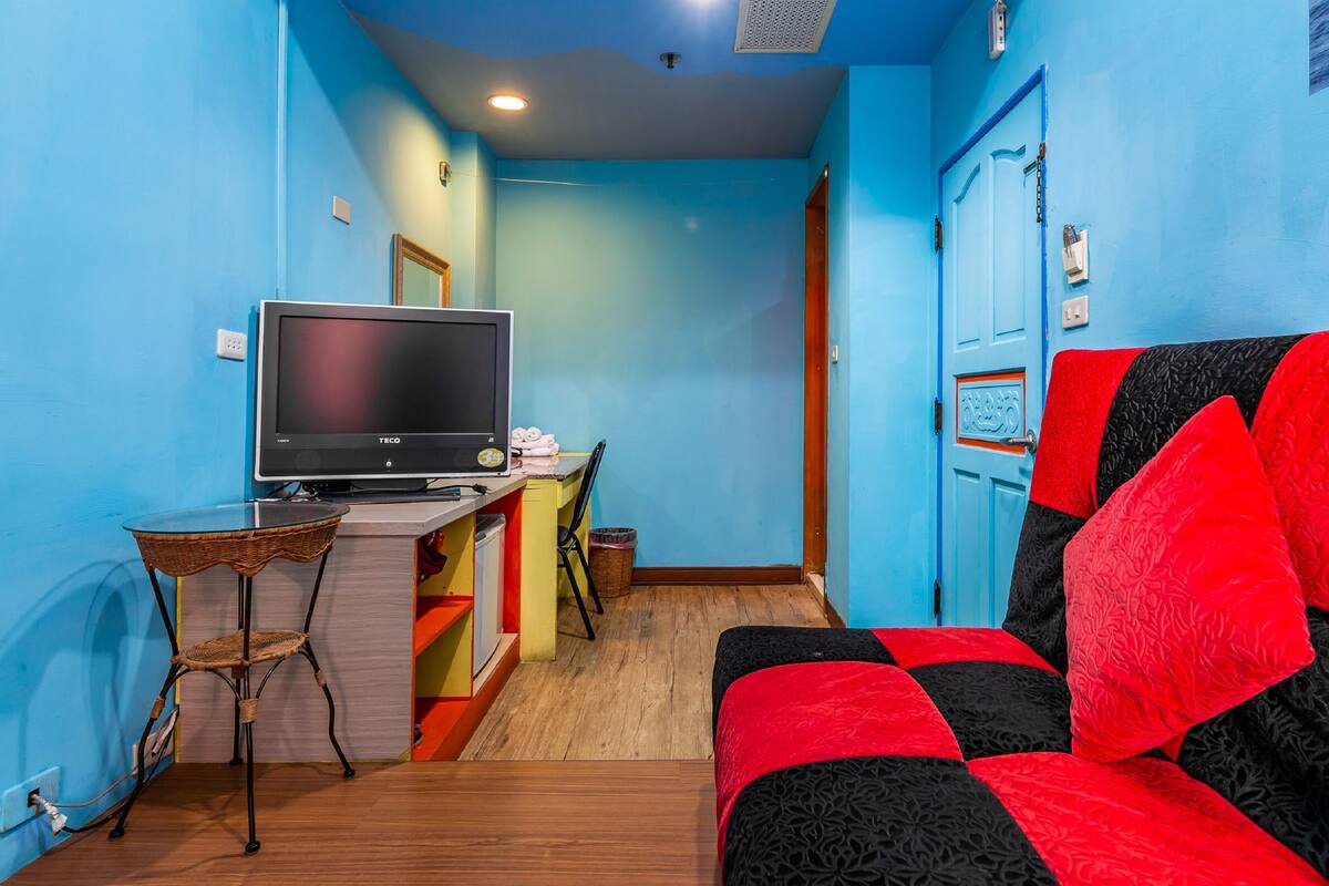 A room with a TV on a stand, a small table with a chair, a sofa with red and black cushions, and a blue door.