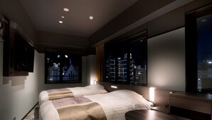 Una habitación de hotel moderna con una cama grande, una mesita de noche, una pequeña mesa redonda y vistas de la ciudad de noche.