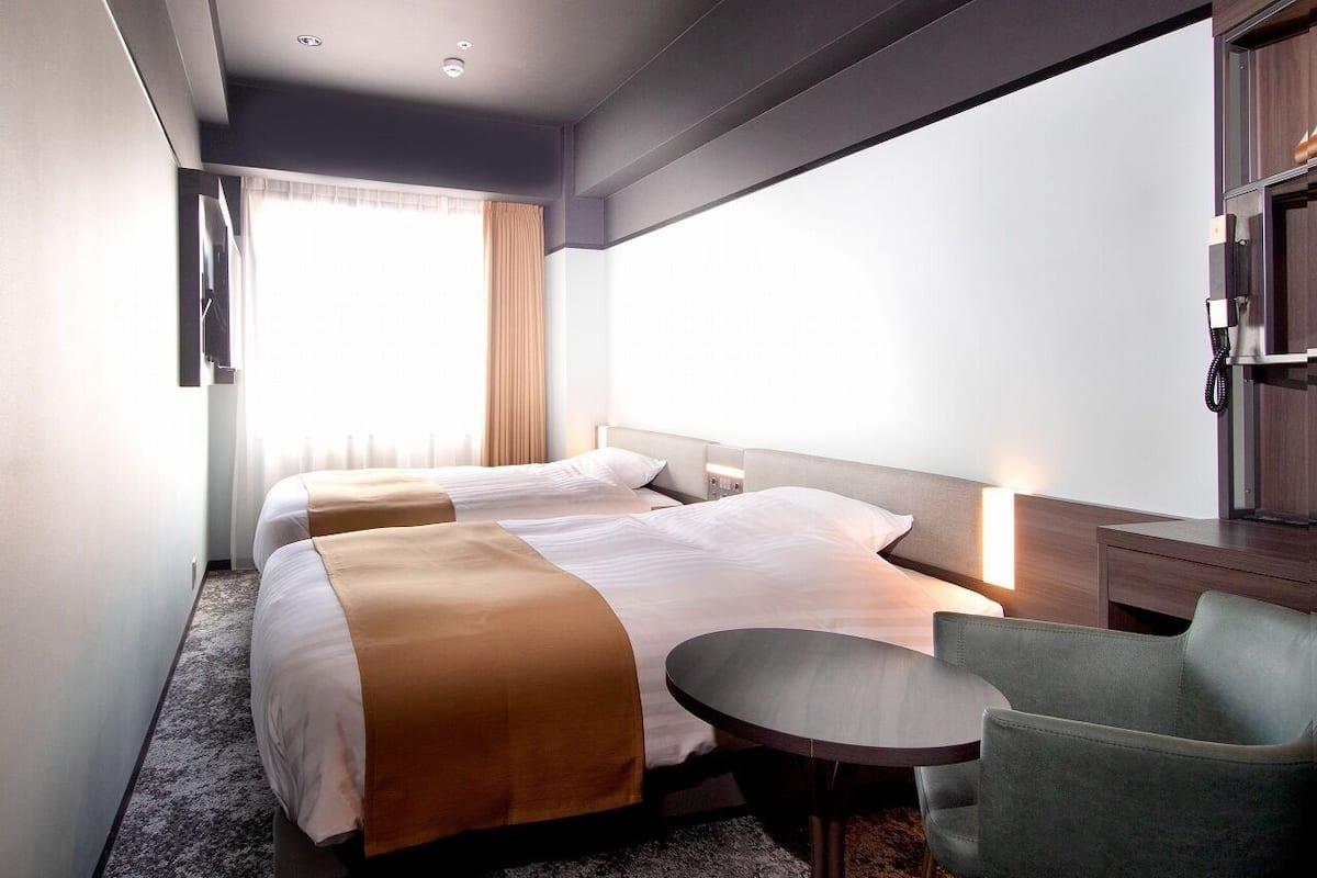 Quarto de hotel com duas camas, uma mesa redonda pequena, uma cadeira, escrivaninha e telefone.
