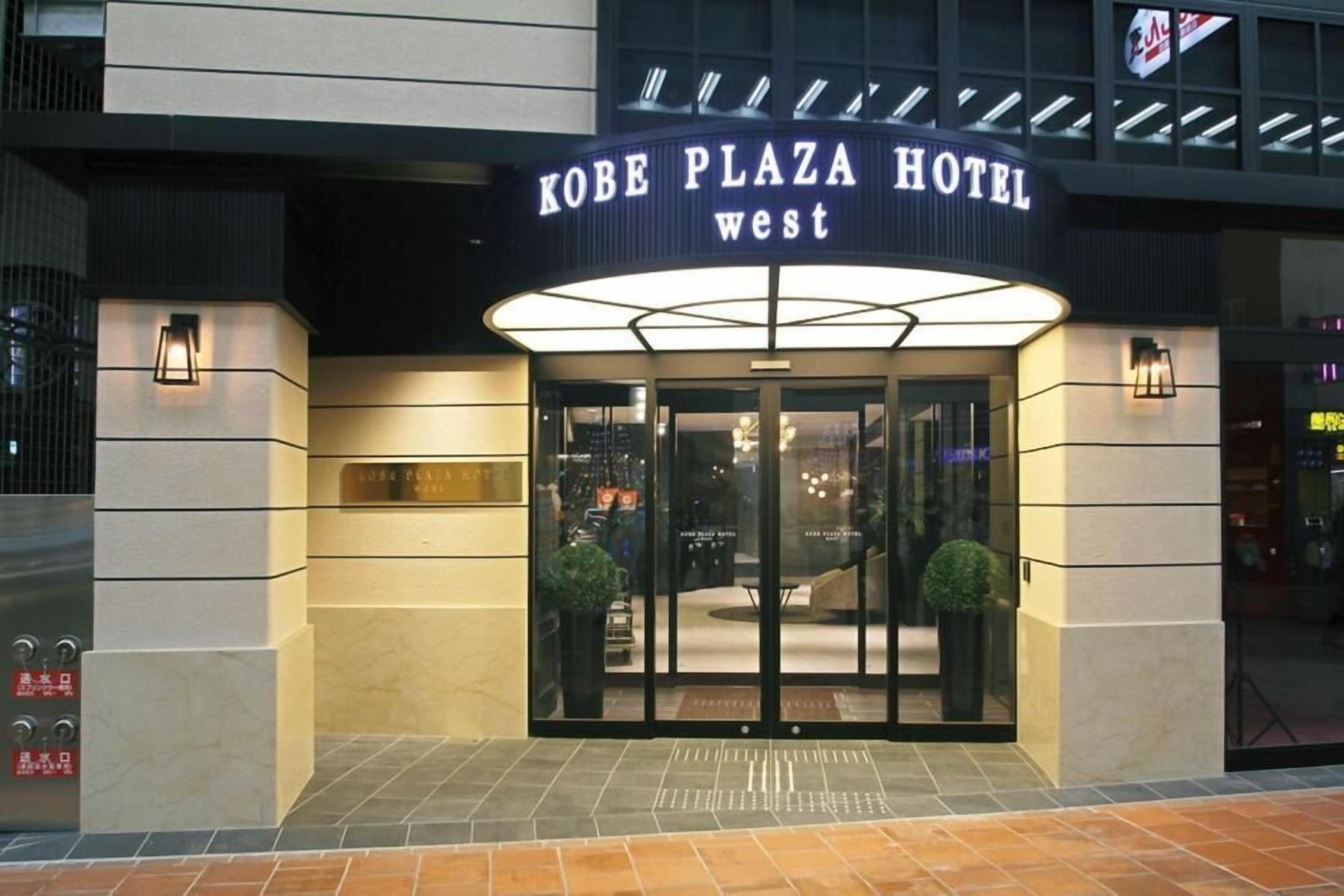 The Kobe Plaza Hotel West entrance with a glass door, a sign above the door, and two potted plants on either side.