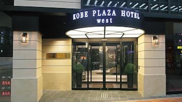 The Kobe Plaza Hotel West entrance with a glass door, a sign above the door, and two potted plants on either side.