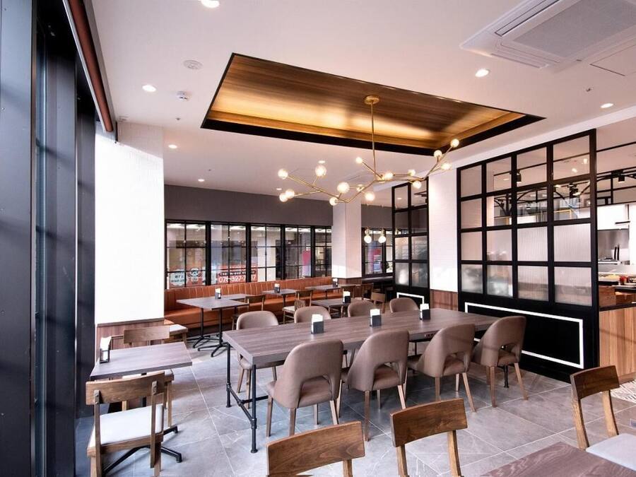 A modern dining area with wooden tables and chairs, a central chandelier, and a kitchen visible through a glass partition.