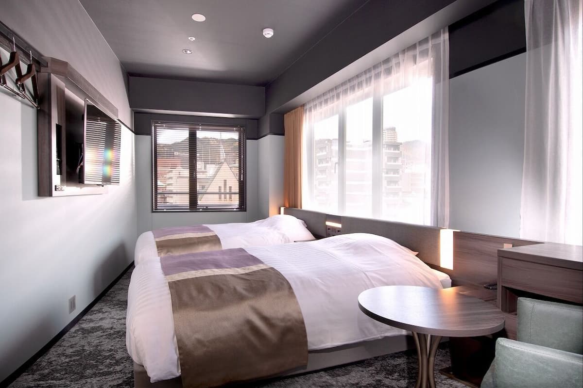 Quarto de hotel com uma cama grande, uma mesinha de cabeceira, uma cadeira, uma janela com persianas e vista para edifícios.
