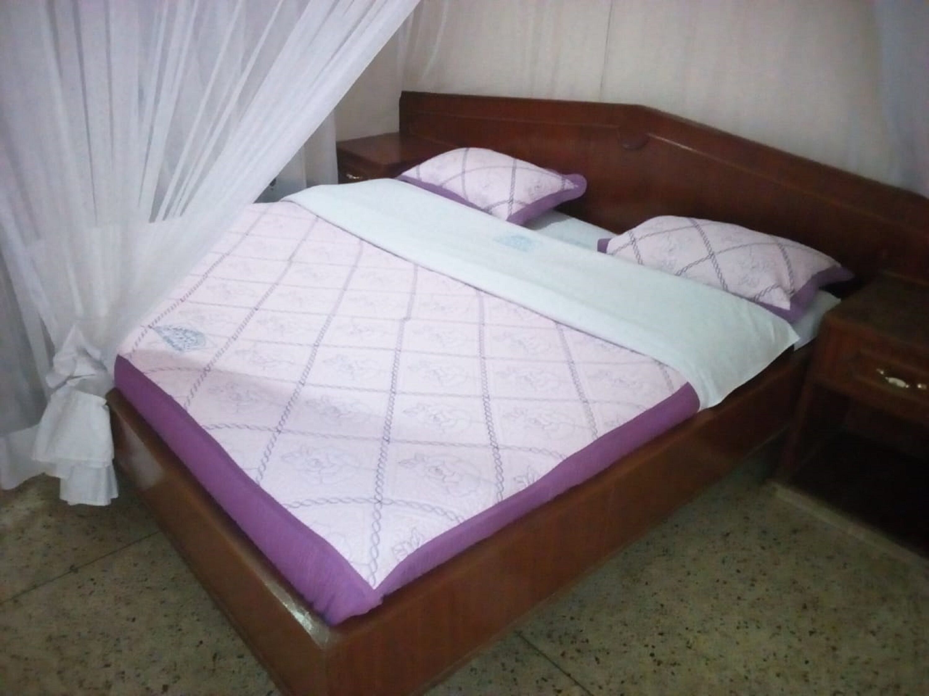 A bed with a wooden headboard, a white and purple bedspread, and a wooden nightstand with a drawer.