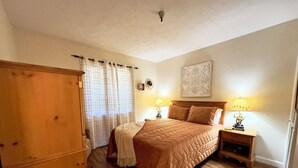 Ein Schlafzimmer mit Bett, zwei Nachttischen mit Lampen, einem Holzschrank, einem Fenster mit Vorhängen und einer Wanddekoration.