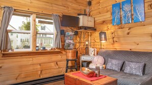 Indretning i en hyggelig hytte med træpaneler, en sofa, et træskab, et vindue med gardiner og dekorationer på væggen.
