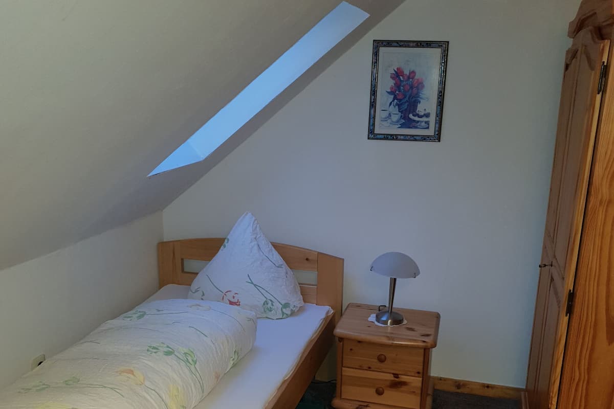 A small, single-bed room with a sloped ceiling, a bedside table with a lamp, and a wooden wardrobe.