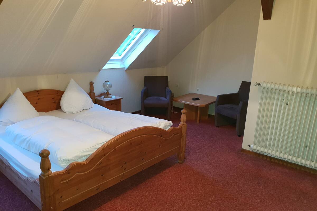 A bedroom with a wooden bed, a small table, a chair, a lamp, and a skylight.