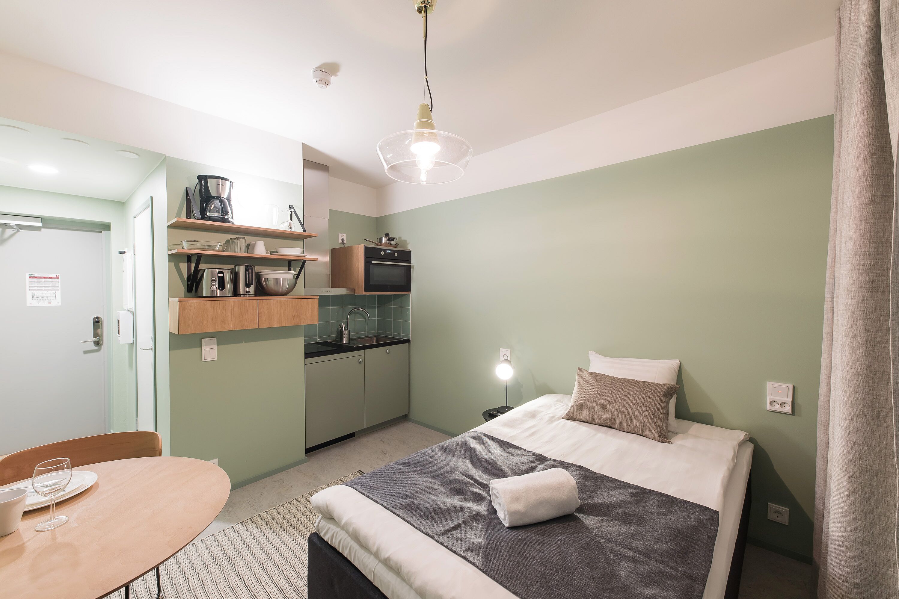 Quarto de hotel pequeno com cama, uma mesinha, cozinha compacta e banheiro.