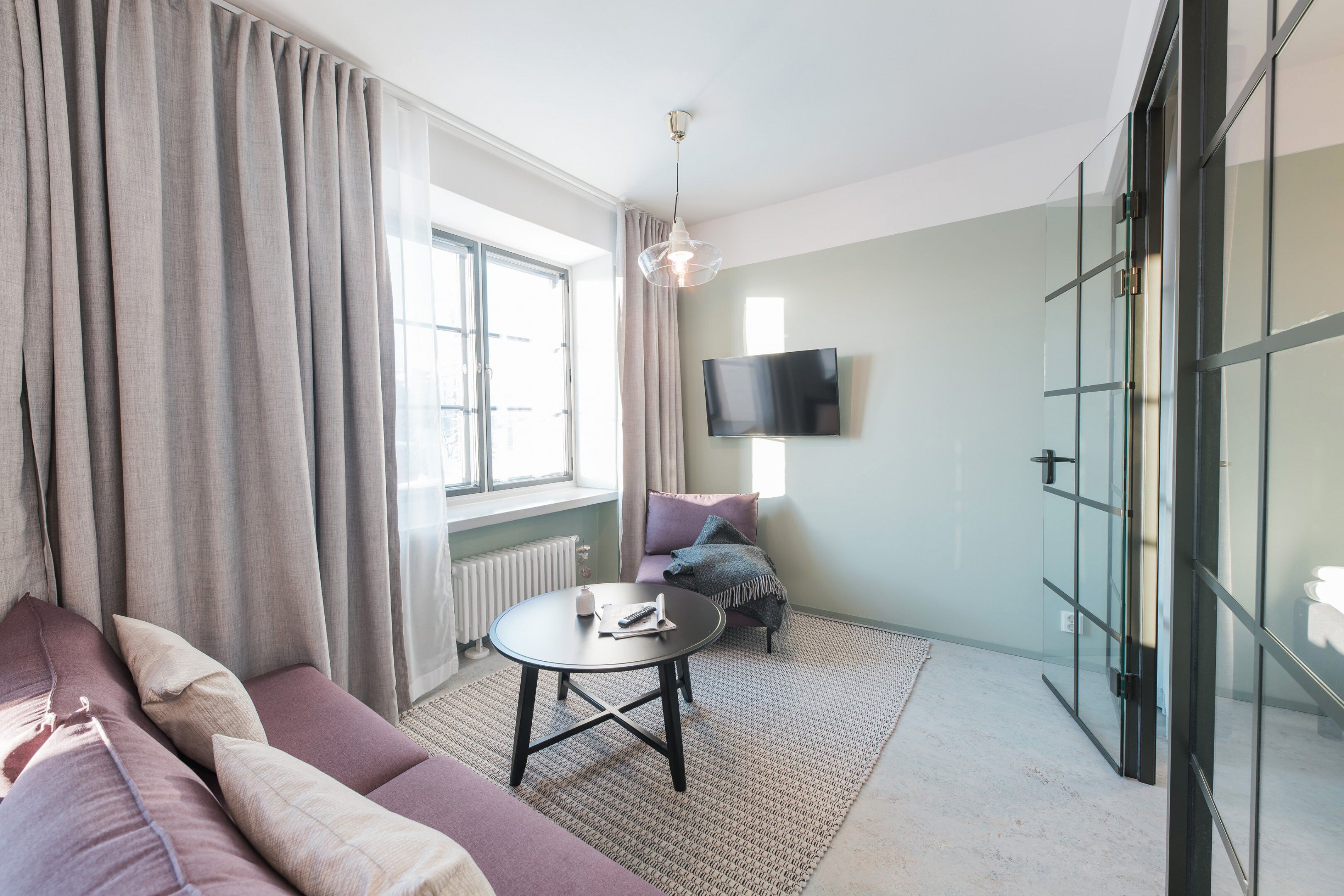 A modern living room with a sofa, a round table, a TV mounted on the wall, and a large window with curtains.