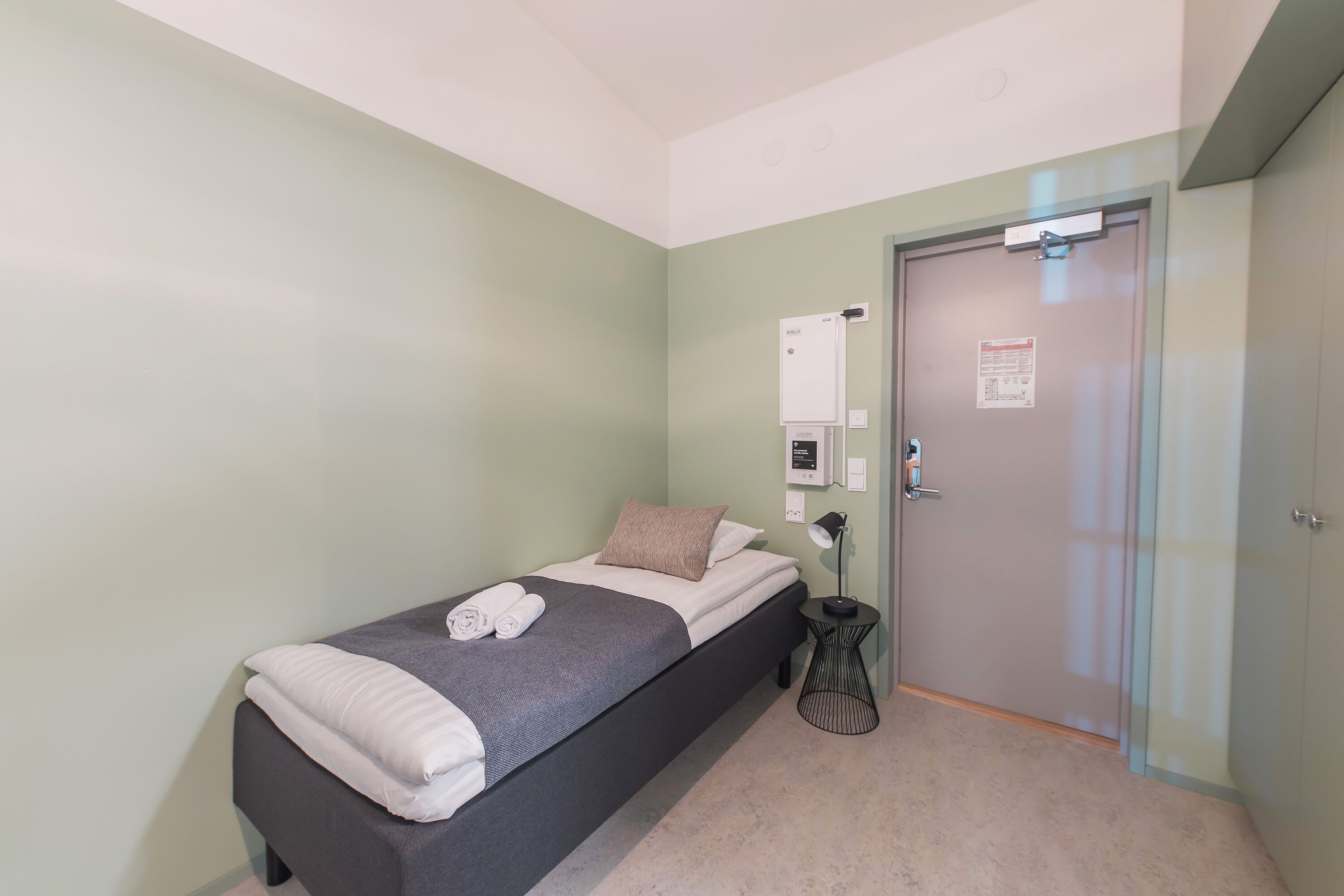 Ein Einzelbett mit grauer Matratze, weißen Kissen und einem gefalteten Handtuch. Ein kleiner Nachttisch mit Lampe und Steckdose ist vorhanden. Die Wände sind hellgrün und die Tür hat ein Milchglasfenster.