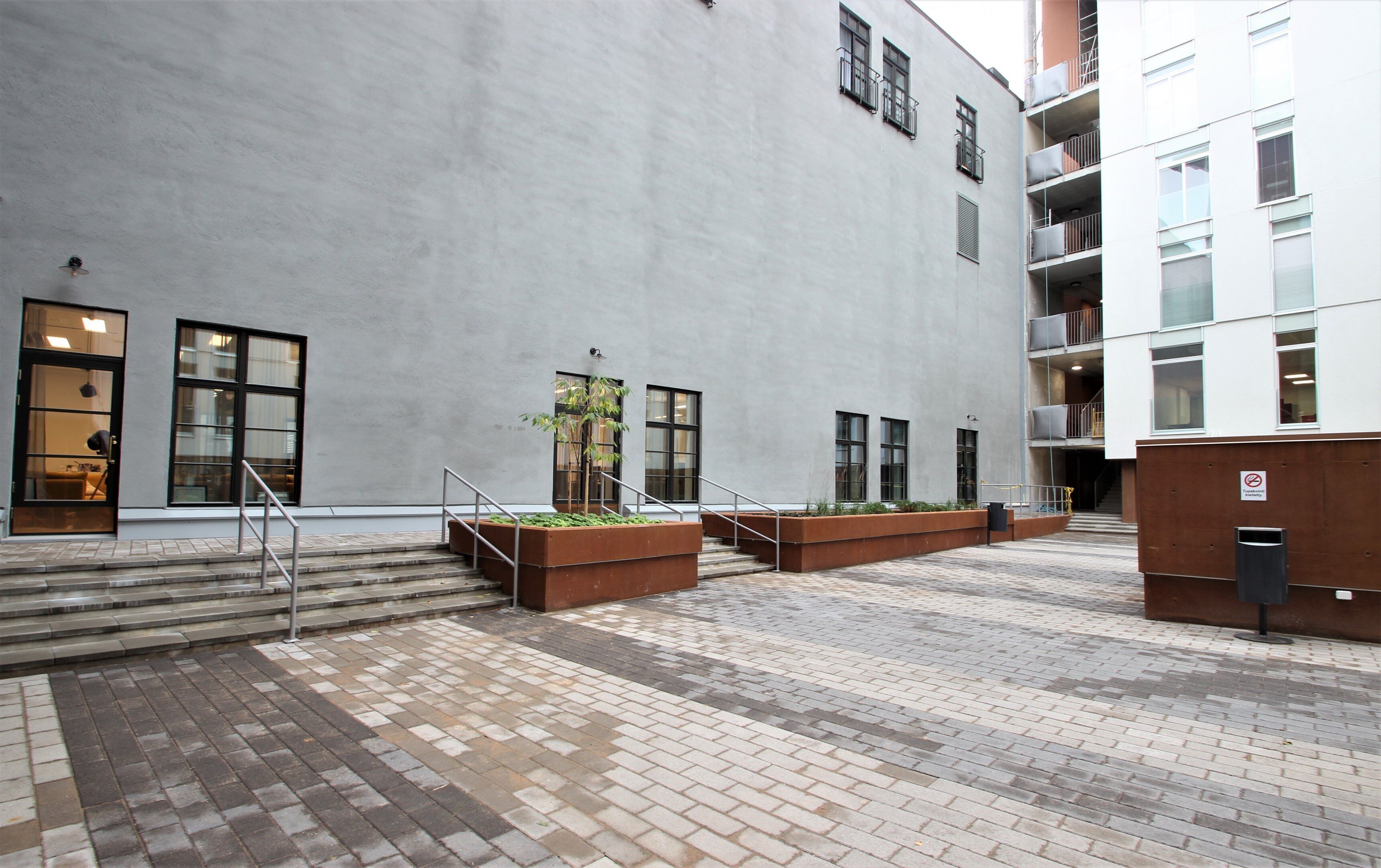 A building with a grey facade, large windows, and a brick-paved courtyard with steps leading to the entrance.