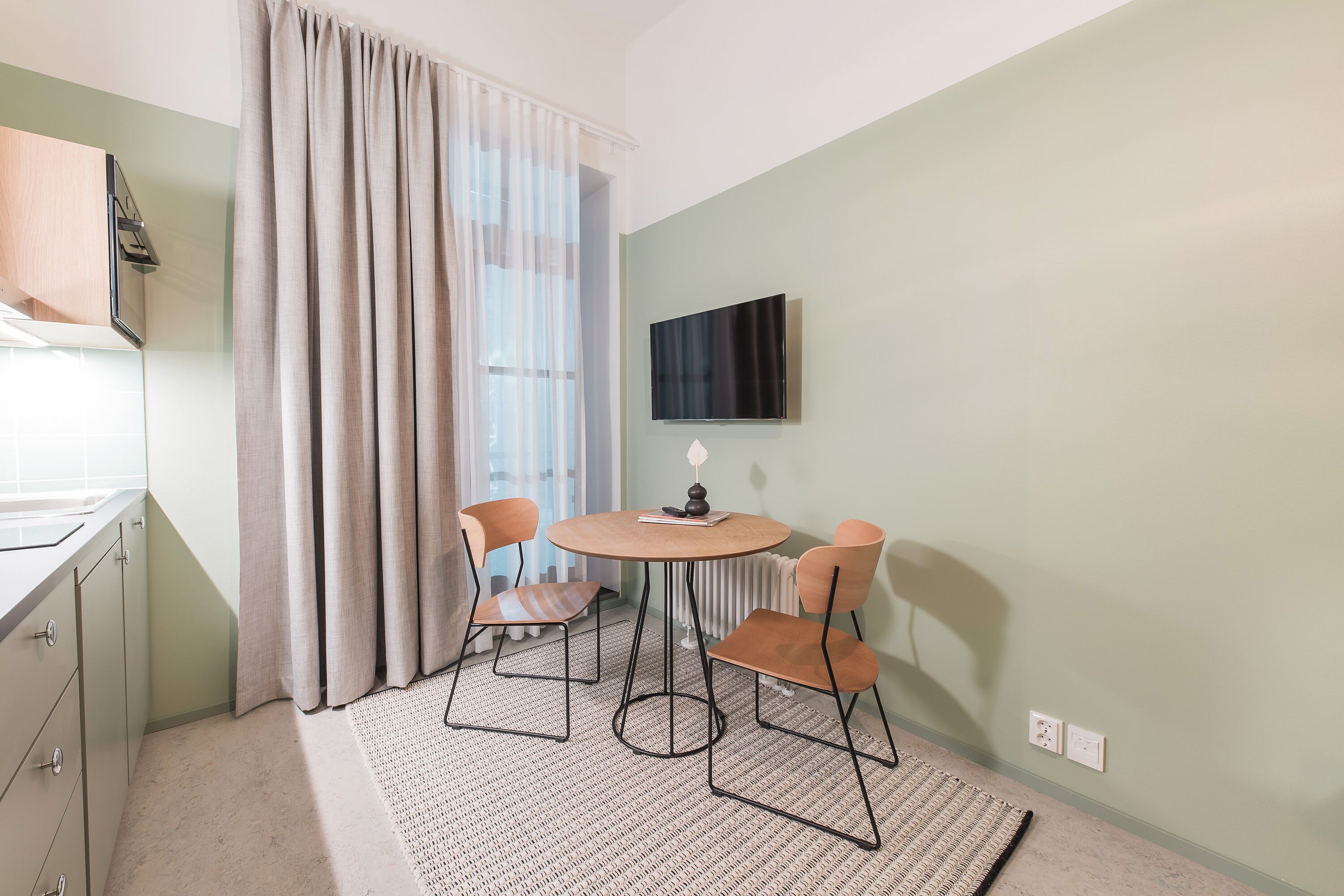 Una habitación pequeña y bien iluminada con una mesa de comedor y sillas, un televisor montado en la pared y una ventana con cortinas.