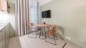 Una habitación pequeña y bien iluminada con una mesa de comedor y sillas, un televisor montado en la pared y una ventana con cortinas.