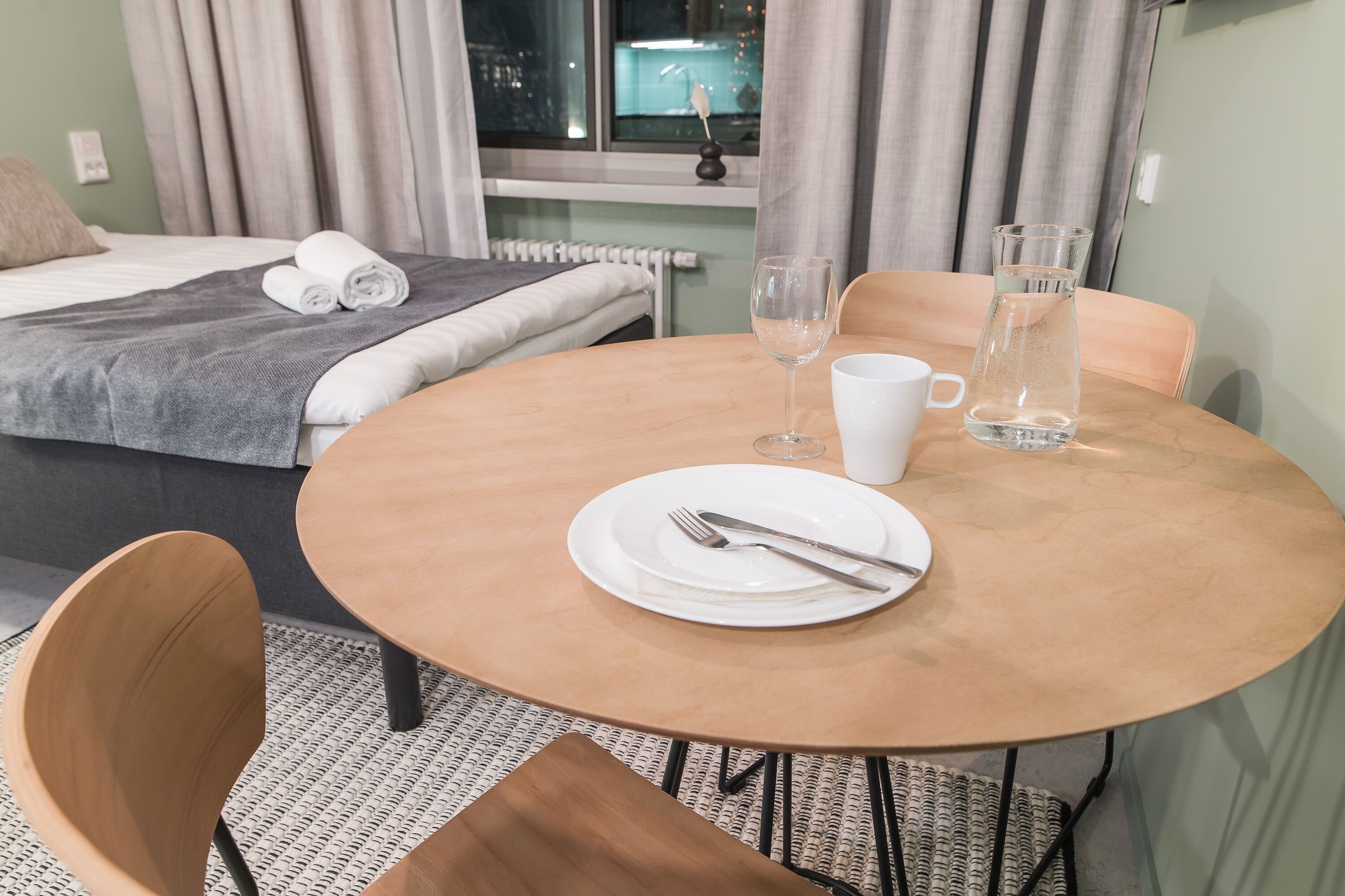 Een keurig opgemaakte hotelkamer met een bed, een ronde tafel met bord, bestek, een glas en een karaf.