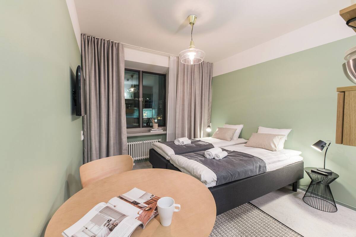 A modern hotel room with a bed, a round table with a magazine and a cup, a TV mounted on the wall, and a window with curtains.