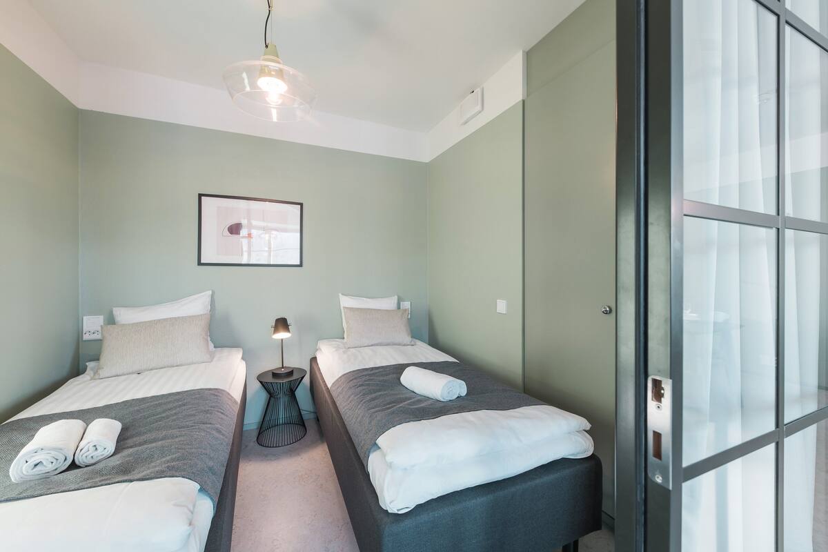 A twin room with two beds, a bedside table with a lamp, a framed picture on the wall, and a sliding glass door.