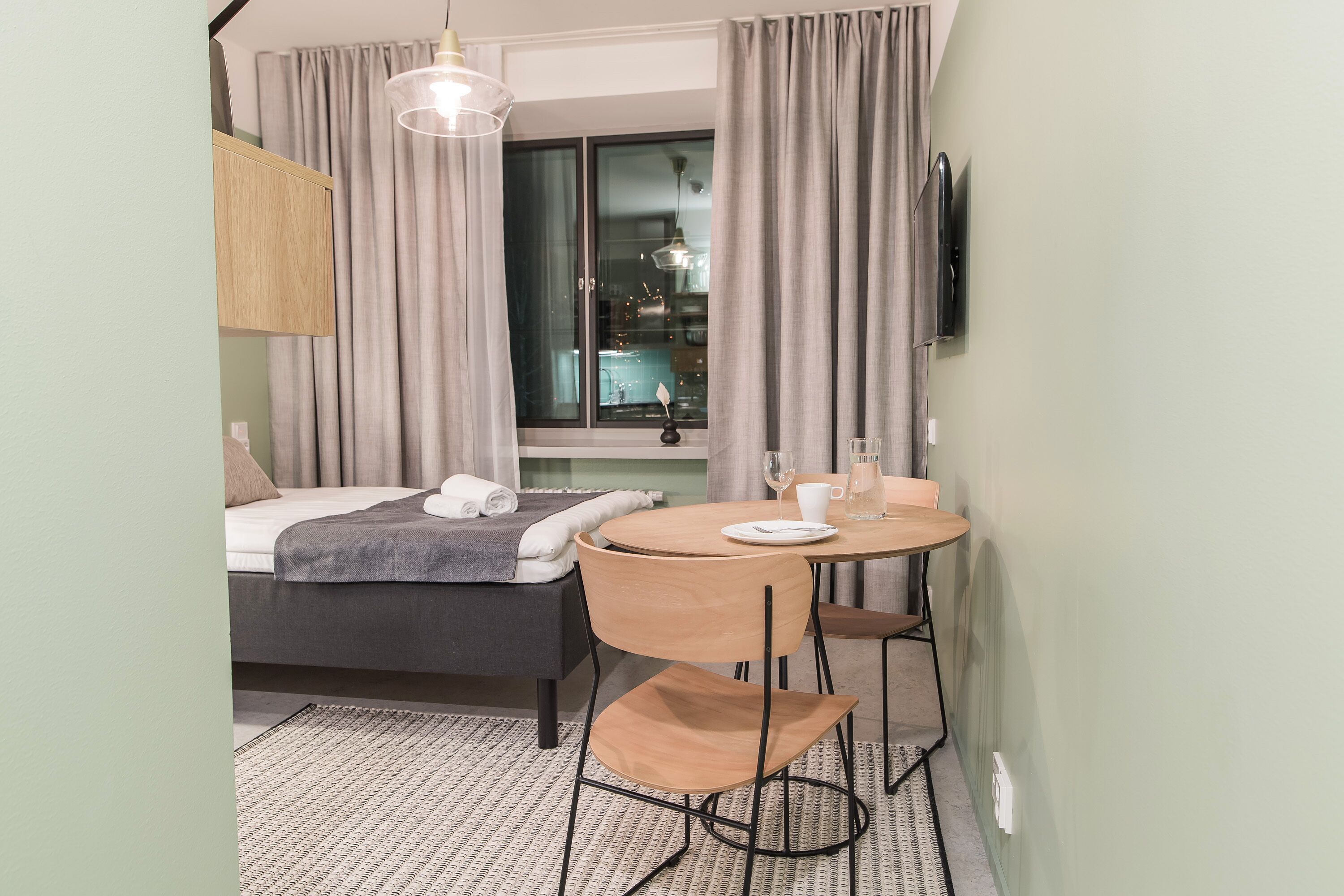 Una habitación de hotel con cama, una mesa de comedor con sillas, una ventana con cortinas, un televisor montado en la pared y una lámpara colgante.
