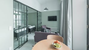 Habitación de hotel moderna con un separador de vidrio que divide la sala de estar del dormitorio. En primer plano, una mesa redonda con un frutero.