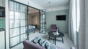 Una habitación de hotel moderna con sofá, una mesita redonda, televisor montado en la pared y una puerta corrediza de vidrio que da al baño.
