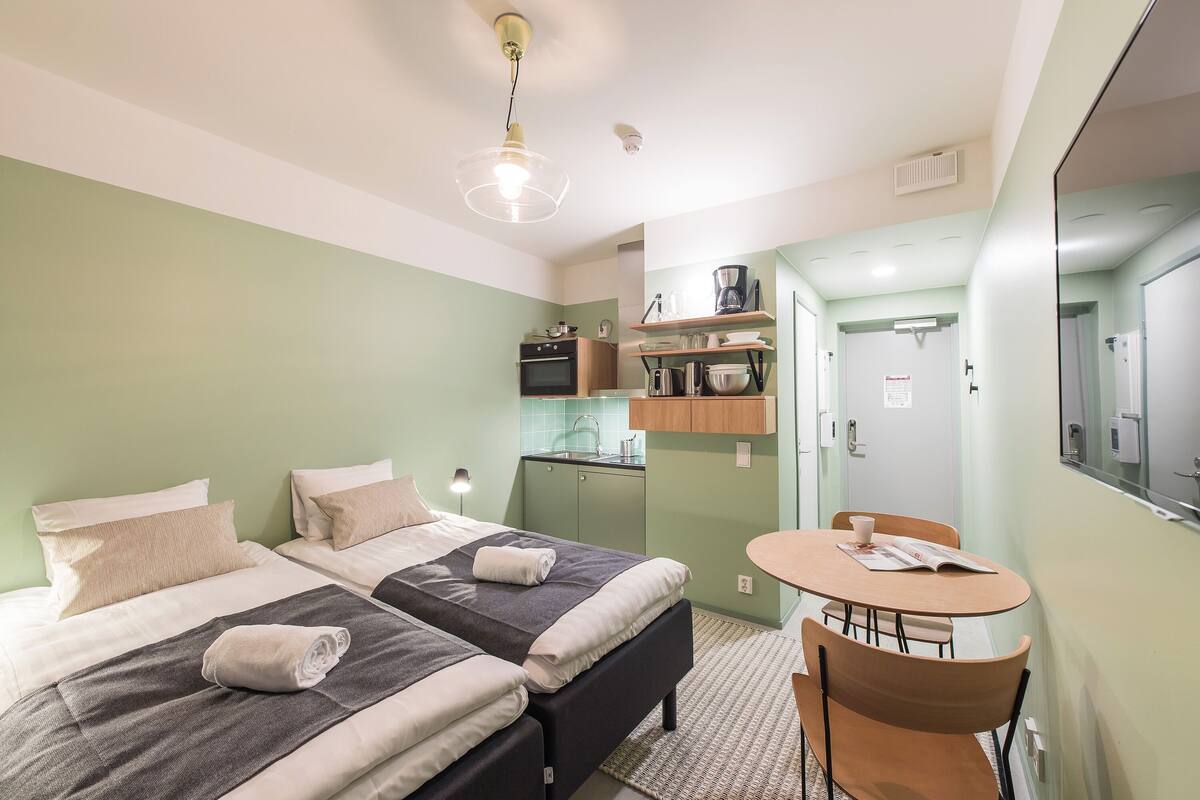 A compact hotel room with two beds, a small kitchenette, a dining area, and a mirror.
