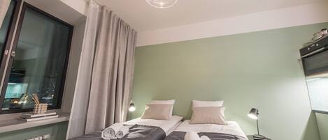 A hotel room with two beds, a window with curtains, a radiator, and a ceiling light fixture.