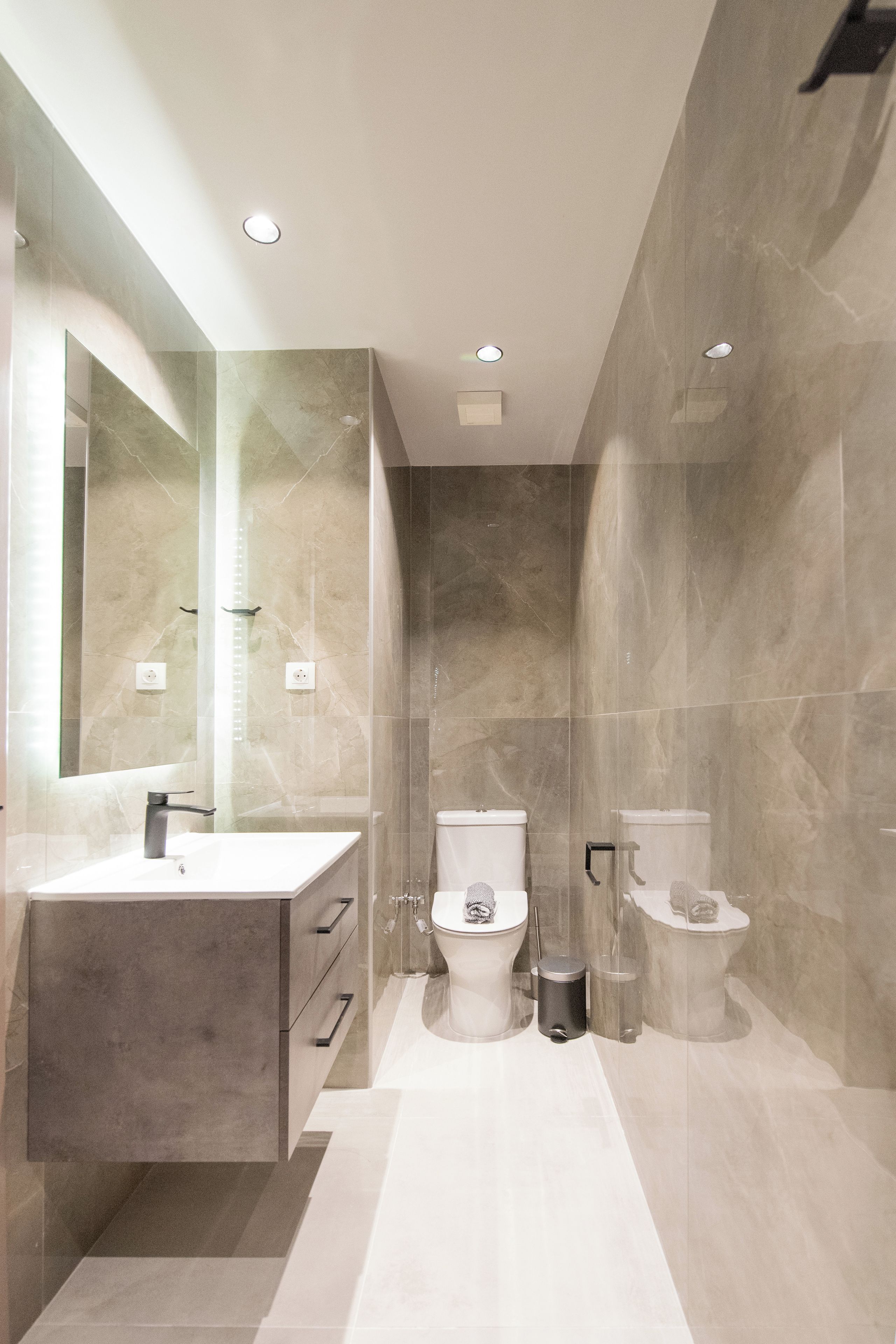 Un baño moderno con un espejo grande, un lavamanos, dos inodoros y un cubo de basura.