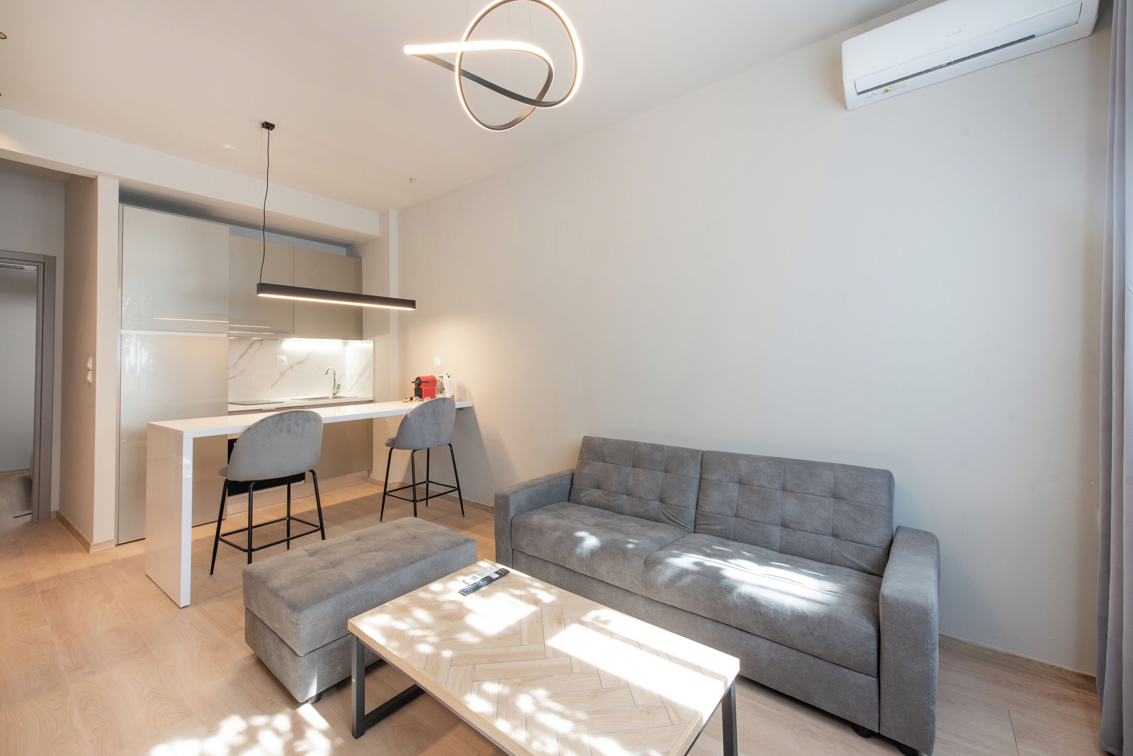A modern living room with a grey sofa, a wooden coffee table, and a kitchen area with a white countertop and chairs.