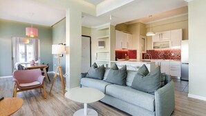 Un salon moderne avec un canapé gris, une petite table ronde et un fauteuil rose. On aperçoit également un coin cuisine avec des meubles blancs et un plan de travail rouge en arrière-plan.