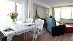 Un salon moderne avec une table à manger pour deux personnes, des chaises en osier blanc, un canapé bleu et un pouf rouge.