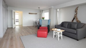 Un salon moderne avec un canapé gris, un pouf rouge et une petite table. Une grande fenêtre avec des rideaux est visible, et une sculpture en bois est accrochée au mur.