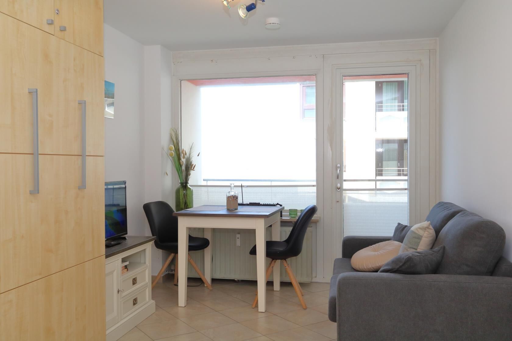 Un salon moderne avec un canapé gris, un meuble en bois, une table à manger avec deux chaises et une grande fenêtre donnant sur l’extérieur.