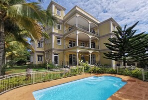 A multi-story building with a swimming pool, surrounded by lush greenery and palm trees.