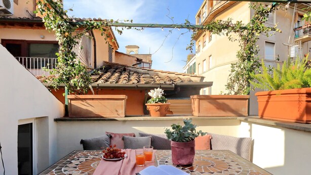 Eine Terrasse mit Tisch für zwei Personen, mit einem Buch, einer Traubenteller und Orangensaftgläsern. Es gibt Blumentöpfe und einen Blick auf ein Gebäude mit einem Ziegeldach.