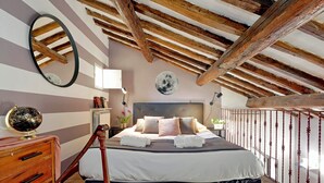 Ein Schlafzimmer mit einem Bett, Holzbalken an der Decke, einem runden Spiegel, einem Globus und einem Nachttisch mit Büchern.
