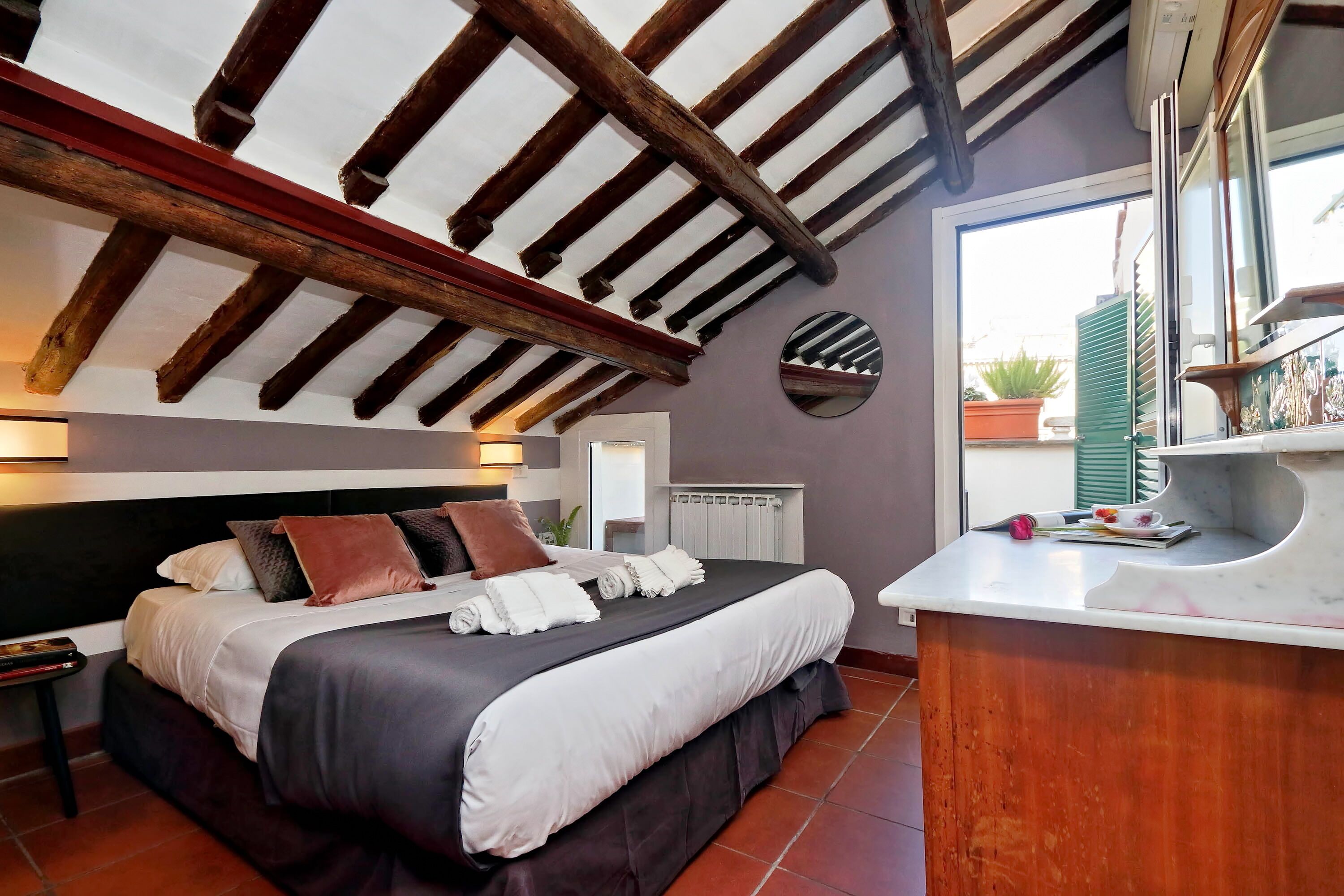 A bedroom with a large bed, wooden beams on the ceiling, a bedside table, and a view of the outside.