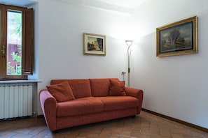 A room with a red sofa, two framed pictures on the wall, a floor lamp, and a window with blinds.