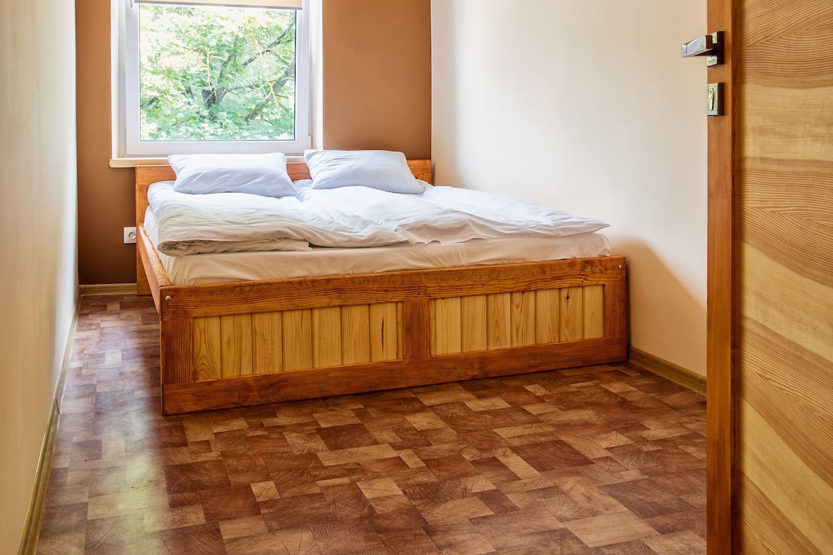 A small bedroom with a wooden bed, a window with a blind, and a wooden door.