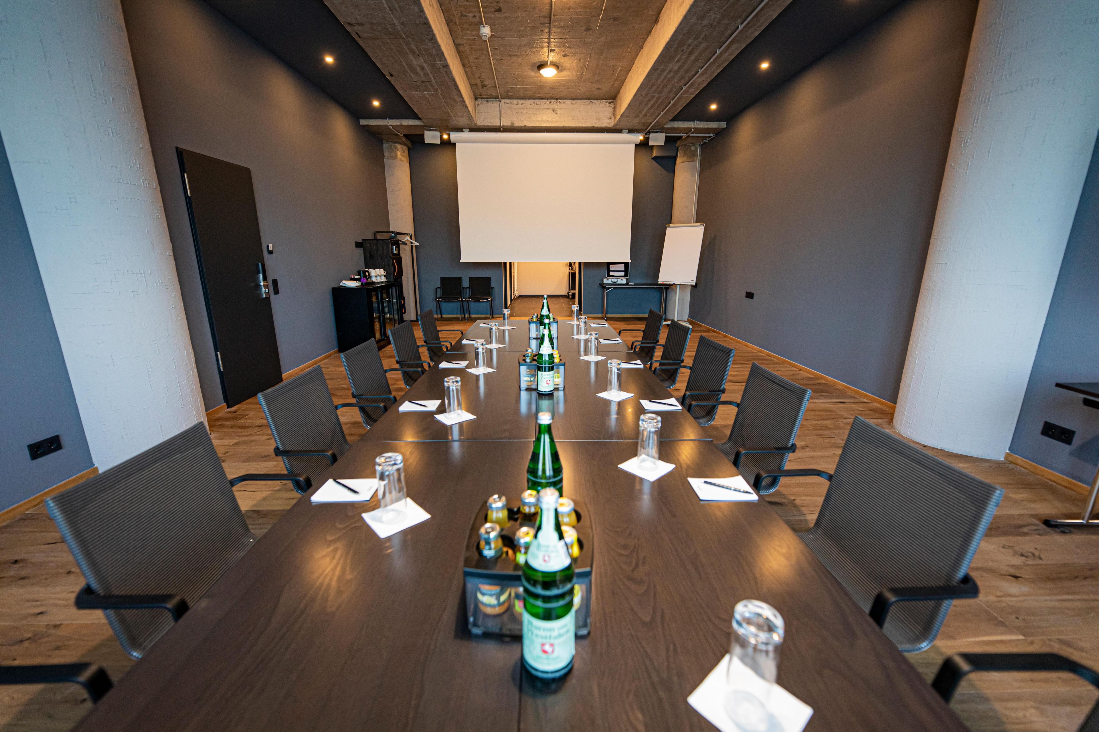 Un salón de conferencias con una larga mesa, sillas, una pantalla de proyector y una pizarra.