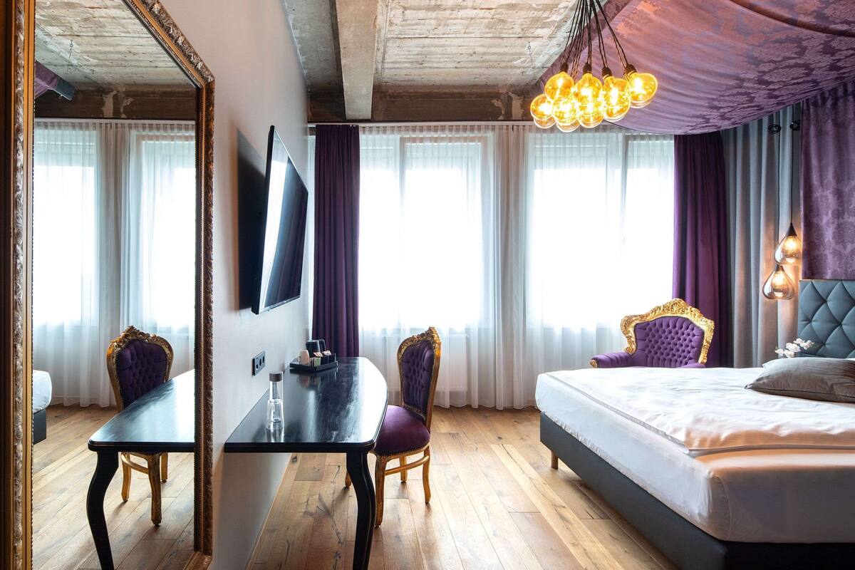 A hotel room with a large bed, a dining table, two chairs, a TV, and a chandelier.