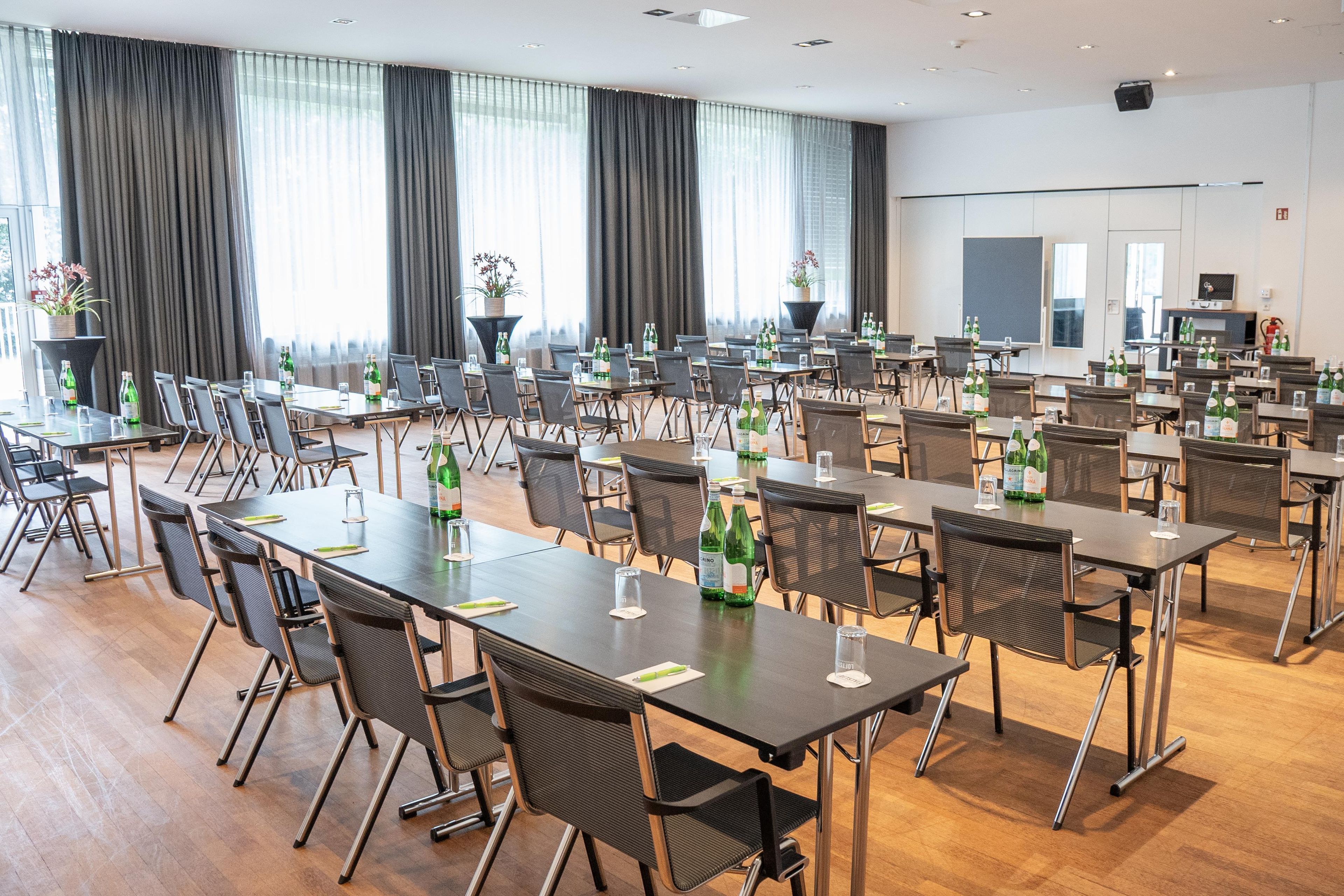 Un salón de conferencias con varias mesas y sillas, ventanas amplias con cortinas y plantas en macetas.