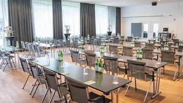 Un salón de conferencias con varias mesas y sillas, ventanas amplias con cortinas y plantas en macetas.