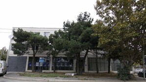 Um edifício moderno com fachada de vidro ampla, área de estacionamento e algumas árvores na frente.