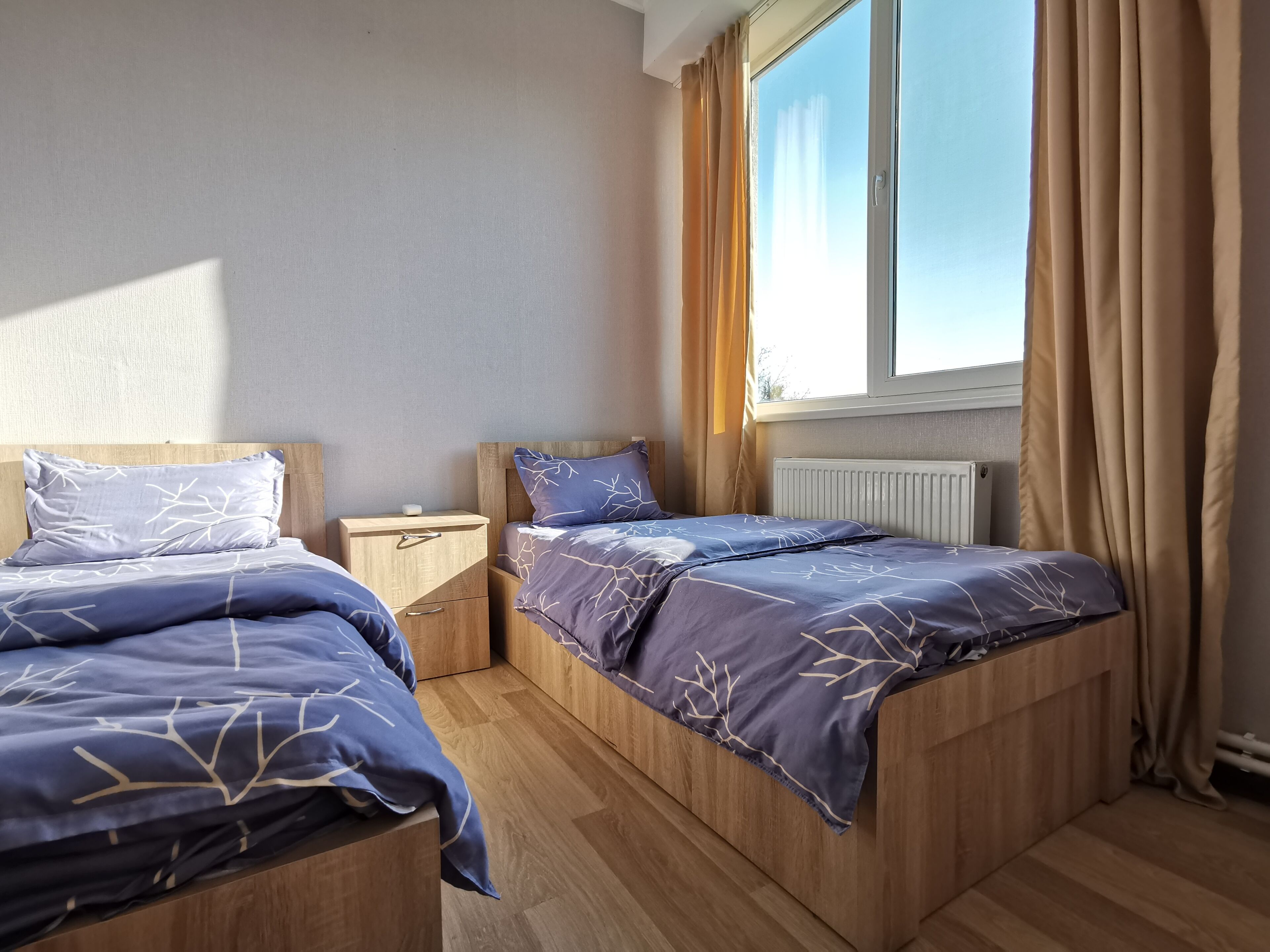 Een slaapkamer met twee bedden, houten hoofdeinden, een nachtkastje, een raam met gordijnen en een radiator.