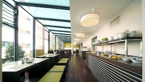 A modern restaurant with long communal tables, a buffet area with plates and bowls, and large windows providing natural light. - Boutiquehotel am Stadtgarten (Freiburg im Breisgau)