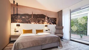 A modern hotel room with a large bed, a nightstand with a lamp, a chair, and a view of a cityscape through a large window. - Boutiquehotel am Stadtgarten (Freiburg im Breisgau)