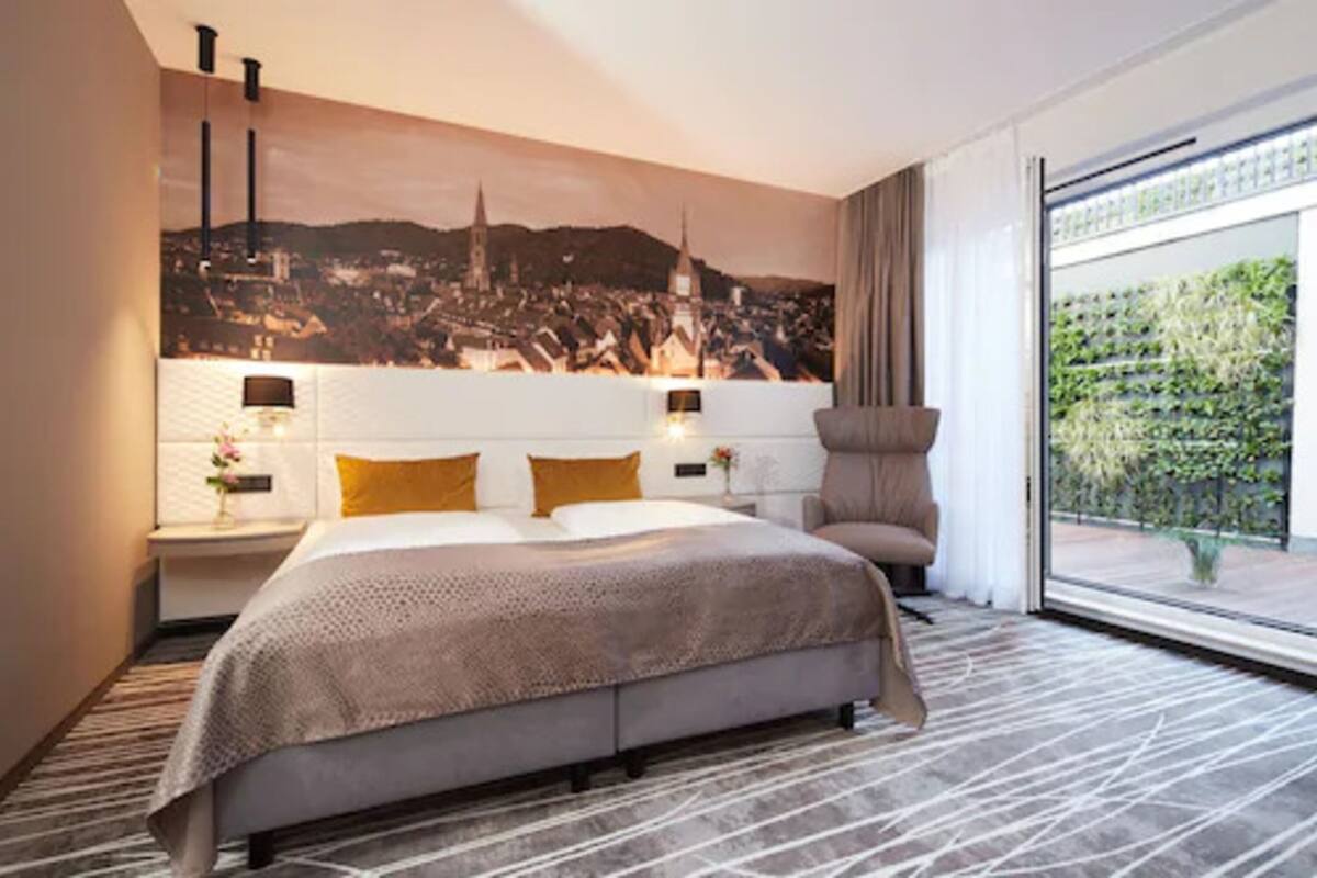 Una habitación de hotel moderna con una cama grande, una mesita de noche con lámpara, una silla y vistas a un paisaje urbano a través de un ventanal amplio.