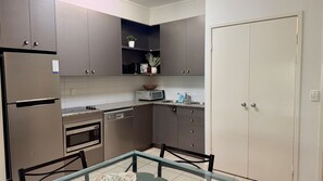 A compact kitchen with a refrigerator, microwave, oven, and a small dining area with chairs and a glass table. - Baden 45 - Rainbow Shores. Beachside Unit for the family. Aircon. Swimming Pool. Tennis Court (Rainbow Beach)