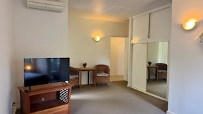 Una camera d'albergo con una TV a schermo piatto su un supporto in legno, una sedia in vimini, un tavolino con una ciotola e una porta scorrevole in vetro.