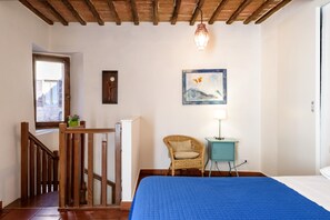 Un dormitorio con una escalera de madera, una cama con colcha azul, una silla de mimbre, una mesita con lámpara y un cuadro en la pared.